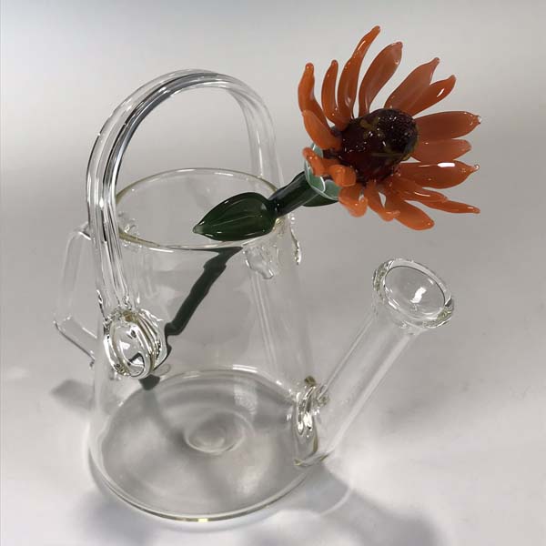 Clear glass watering can. Inside of it, a glass flower with orange petals and a long green stem with two leaves.